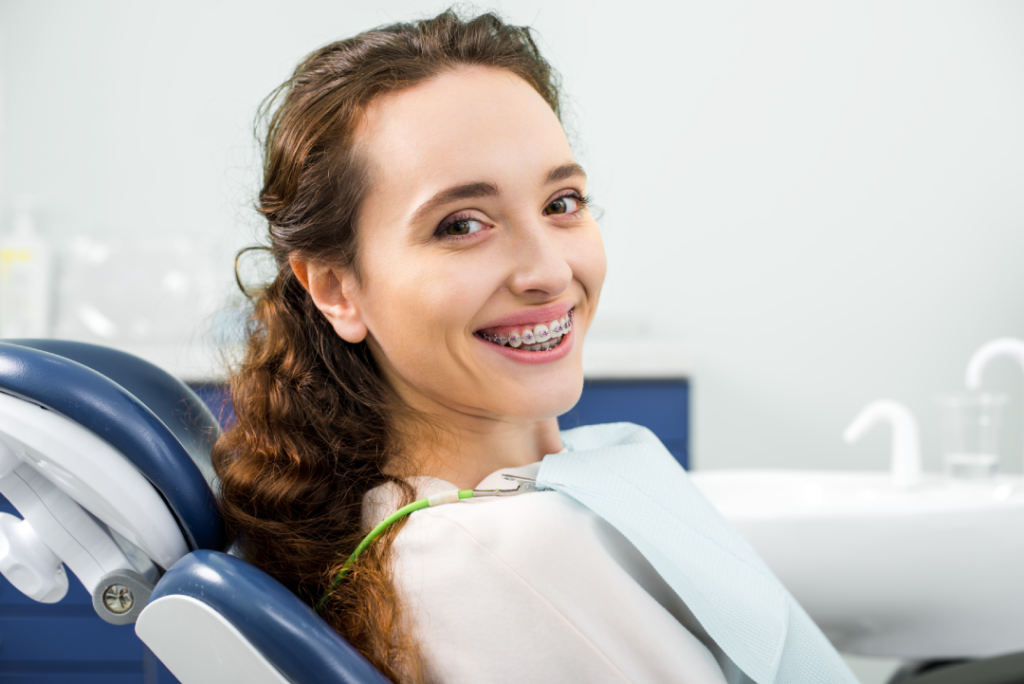 Une jeune femme aux cheveux bouclés et portant un appareil dentaire sourit, assise dans un fauteuil de dentiste, vêtue d'un bavoir de protection, dans un cabinet dentaire lumineux spécialisé dans les bagues dentaires pour adultes à Casablanca et l'orthodontie invisible à Casablanca.