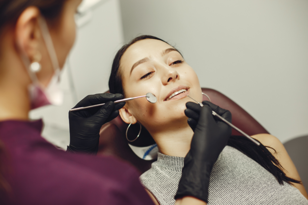 Une femme est allongée dans un fauteuil dentaire, souriante, tandis qu'un dentiste portant des gants noirs examine ses dents à l'aide d'instruments dentaires, illustrant ainsi les résultats spectaculaires des facettes dentaires Casablanca.