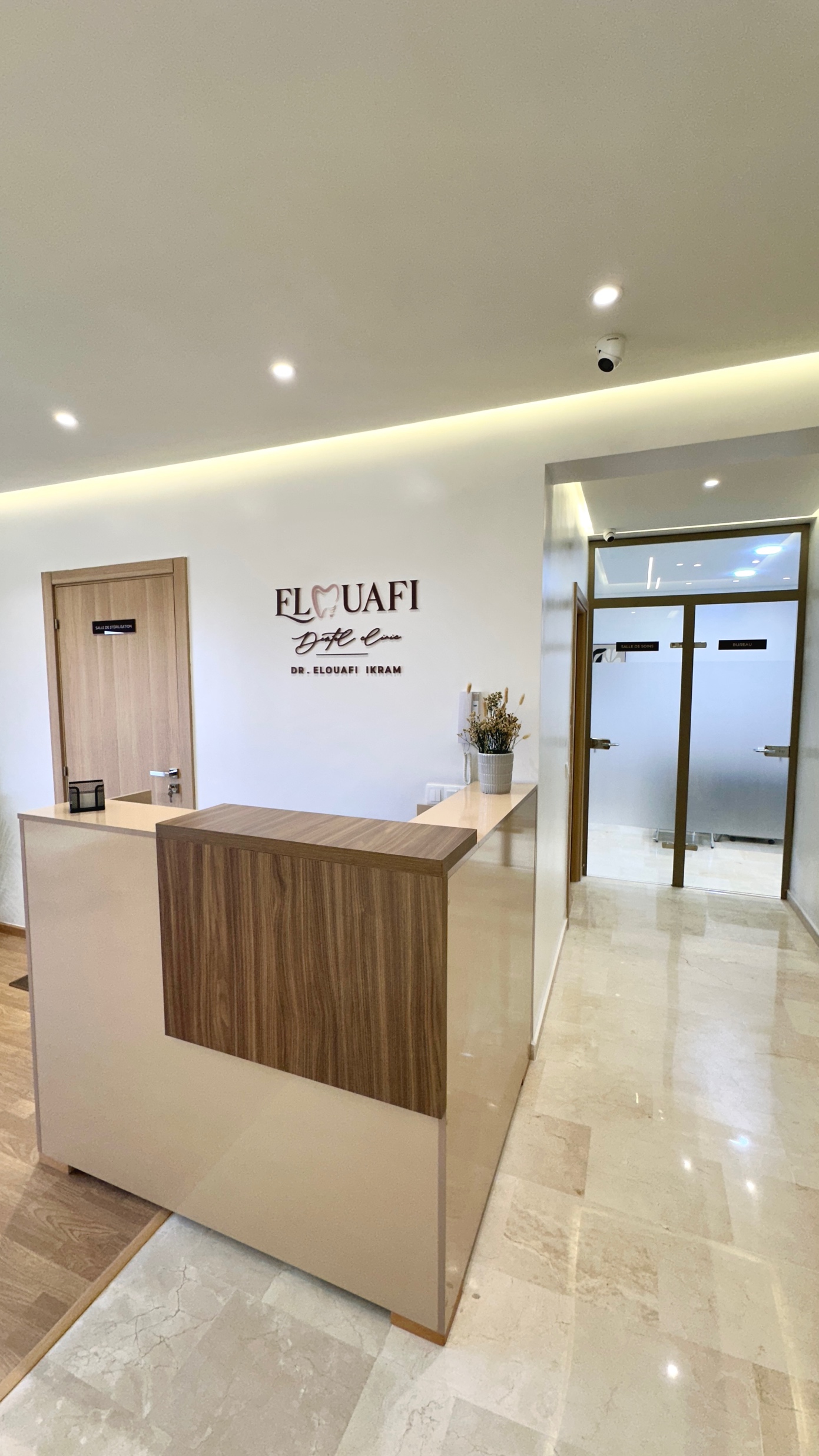 Le hall d'accueil d'un cabinet médical moderne, avec un comptoir en bois, des murs et un sol beiges, une porte portant l'inscription « Dr Elouafi » et des portes en verre dépoli donnant sur le couloir. L'enseigne accrochée au mur indique : « ELOUAFI Dr Ikram Elouafi, dentiste à Casablanca ».