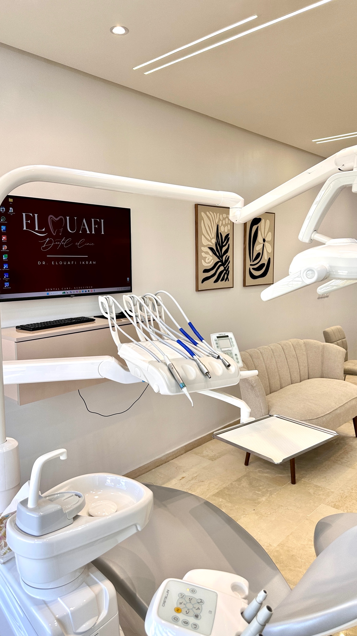 Un cabinet dentaire moderne équipé d'un fauteuil dentaire, d'instruments, d'un canapé beige, de tableaux abstraits aux murs et d'un écran affichant le nom « Elouafi Dente Care ». La pièce est lumineuse, décorée dans des tons neutres et dans un style minimaliste : un espace accueillant, idéal pour ceux qui recherchent un cabinet dentaire à Casablanca ou un dentiste expérimenté à Casablanca.